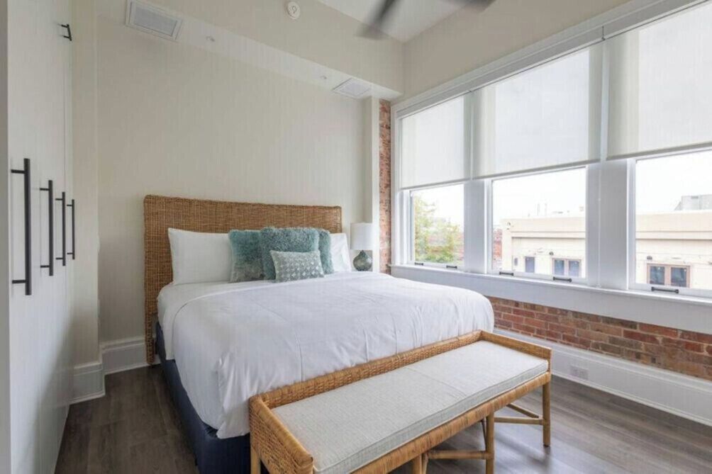 Une chambre avec une grande fenêtre, un lit avec une literie blanche, un banc et un mur en briques.