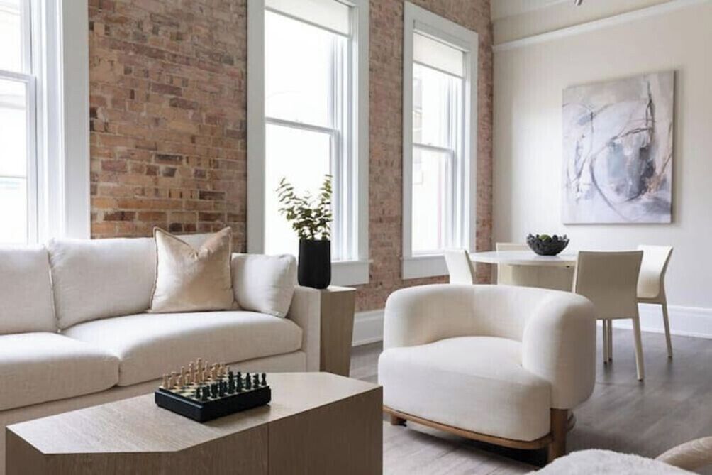 Un salon moderne avec un mur en briques, un canapé, une table basse avec un jeu d’échecs, un coin repas avec une table et des chaises, et un tableau accroché au mur.