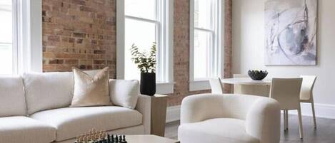 A modern living room with a brick wall, a sofa, a coffee table with chess pieces, a dining area with a table and chairs, and a painting on the wall.