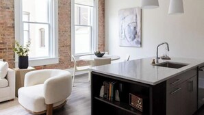 A modern kitchen with a large island, white cabinetry, and a brick wall. - Cotton, Silk, and Linen at the Brent Lofts (Pensacola)
