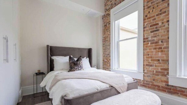 A bedroom with a large window, a brick wall, a bed with a headboard, and a bedside table. - Cotton, Silk, and Linen at the Brent Lofts (Pensacola)