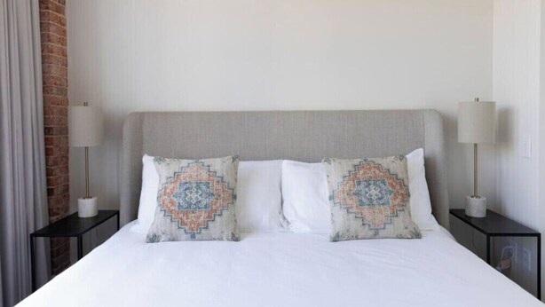 A hotel room with a bed, two nightstands, a lamp, and a brick wall. - 2BR Palafox Oasis at the Brent Lofts (Pensacola)