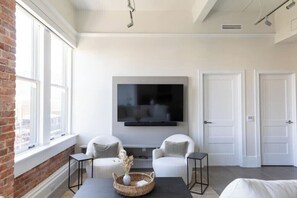 A modern living room with a flat-screen TV mounted on the wall, two white armchairs, a coffee table with a decorative basket, and a brick wall on one side. - 2BR Palafox Oasis at the Brent Lofts (Pensacola)