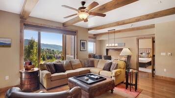 A spacious living room with a large sectional sofa, a leather armchair, and a coffee table. There is a wooden ceiling fan, a large window offering a view of the outdoors, and a door leading to another room.