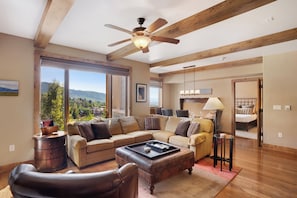 A spacious living room with a large sectional sofa, a leather armchair, and a coffee table. There is a wooden ceiling fan, a large window offering a view of the outdoors, and a door leading to another room. - Edgemont Condominiums (Steamboat Springs)