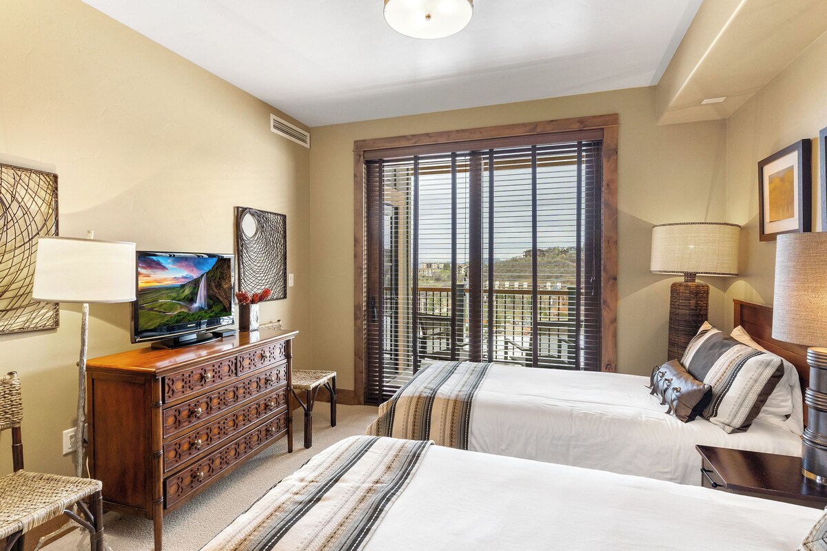 Habitación de hotel con dos camas, televisor, cómoda y ventana con persianas.
