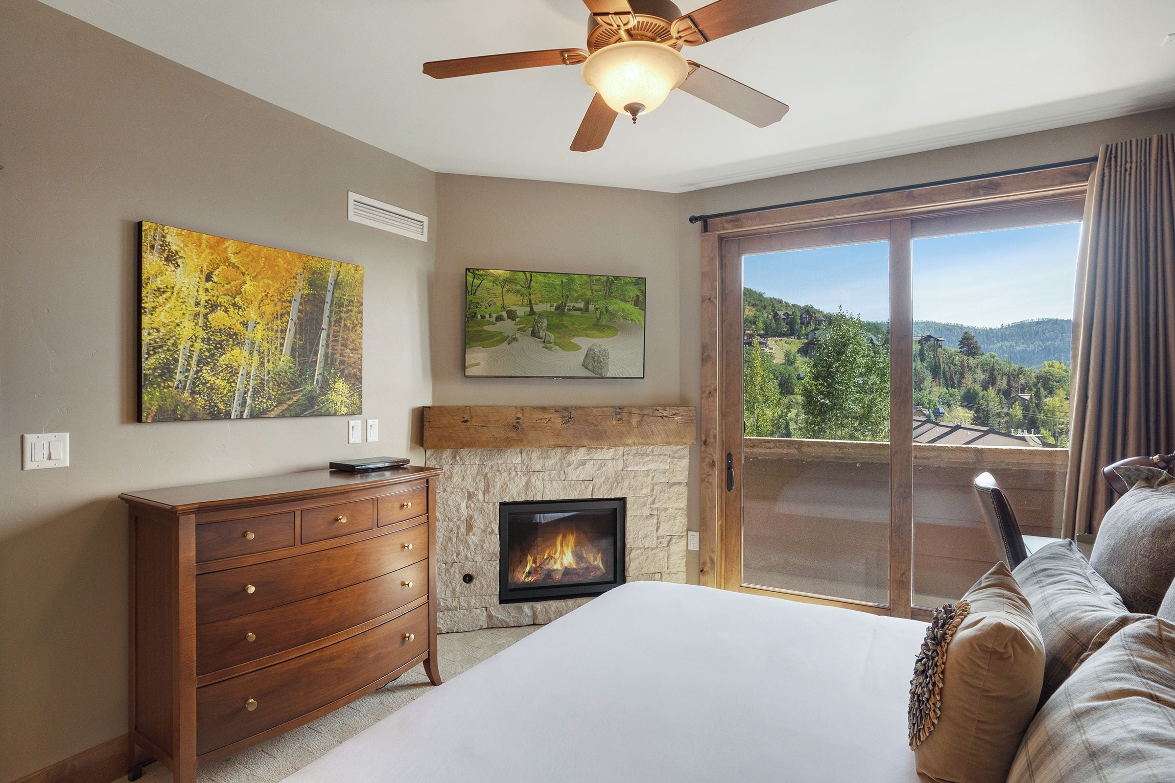 A bedroom with a fireplace, a ceiling fan, a large window with a view, and a wooden dresser.