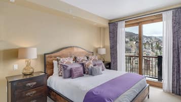 Una camera d'albergo con un letto grande, una testiera in legno, un comodino, una lampada e vista su una zona montuosa dalla finestra.