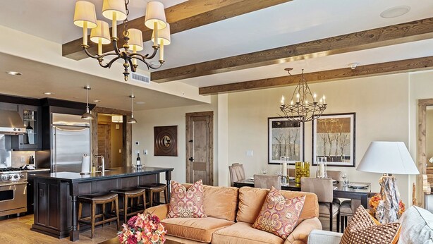 A spacious living area with a large sofa, a coffee table with flowers, a chandelier, and a kitchen in the background. - Edgemont Condominiums (Steamboat Springs)