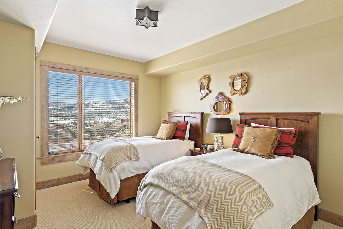 A bedroom with two beds, a large window with blinds, and a view of a cityscape.