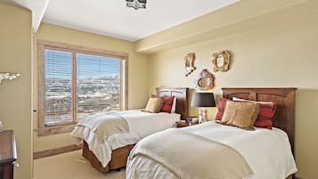 A bedroom with two beds, a large window with blinds, and a view of a cityscape.