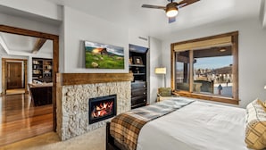 A bedroom with a fireplace, a bed, a television, and a large window with a view. - Edgemont Condominiums (Steamboat Springs)
