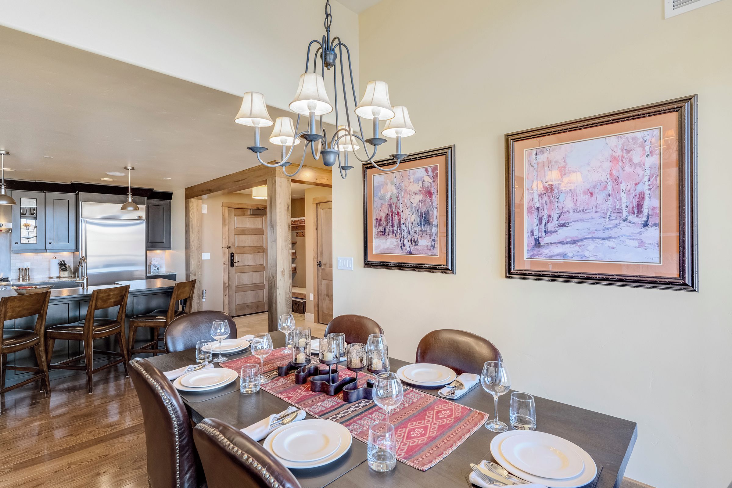 A dining area with a long table set for six, a chandelier, and two framed paintings on the wall.