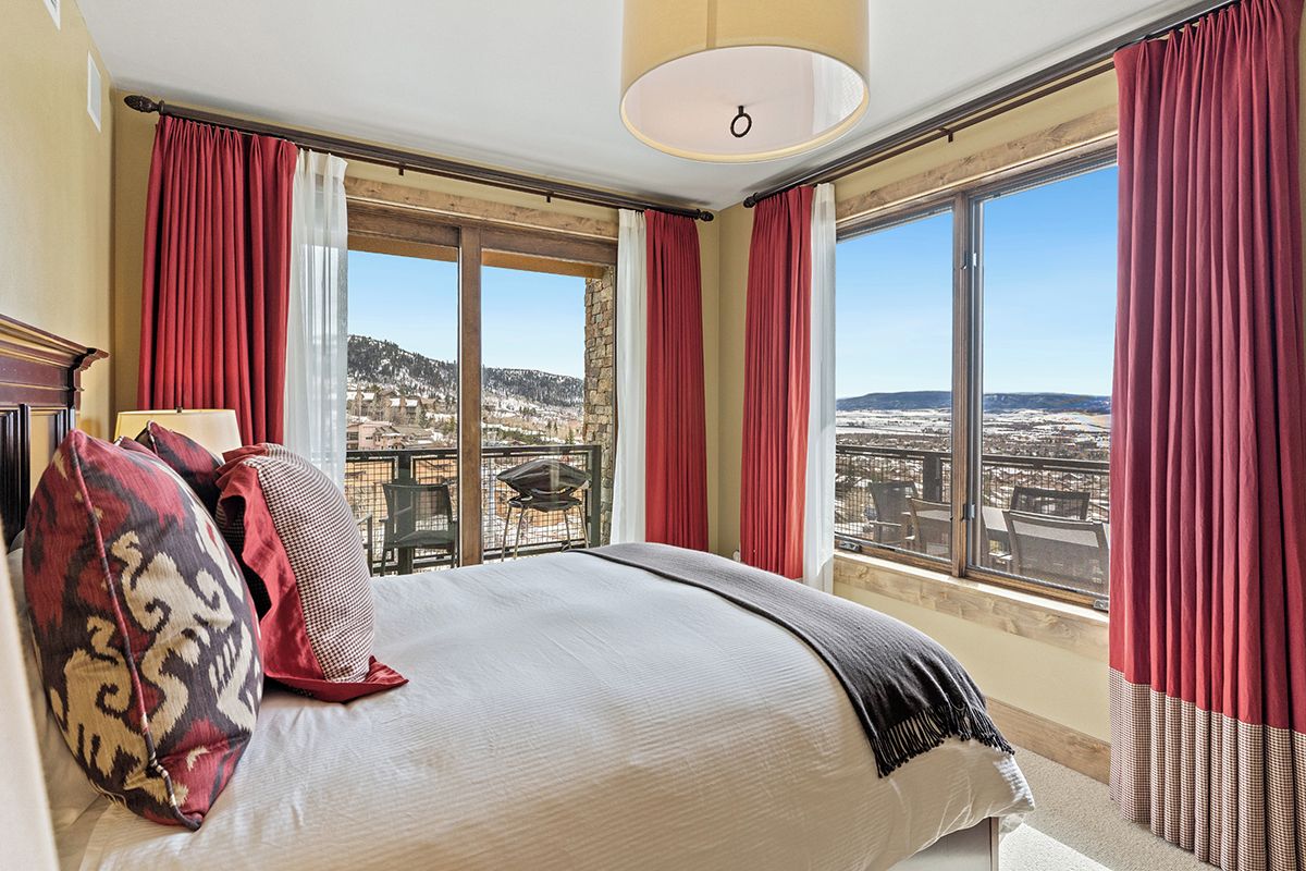 A bedroom with a large bed, red curtains, and a view of a cityscape through large windows.