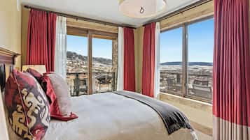 Un dormitorio con una cama grande, cortinas rojas y vistas a un paisaje urbano a través de amplias ventanas.