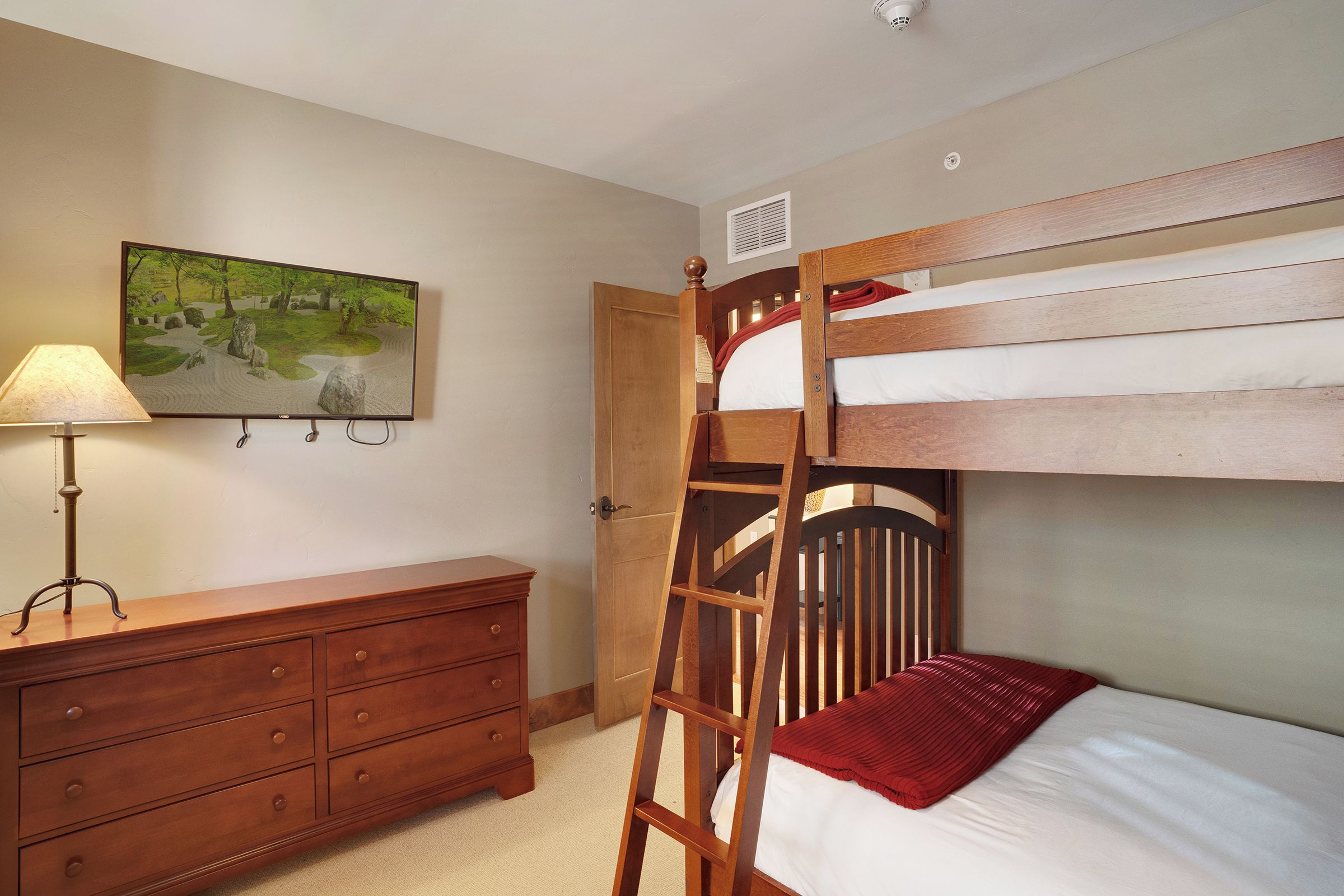 Un lit superposé avec une échelle, une commode en bois, une lampe et un tableau accroché au mur.