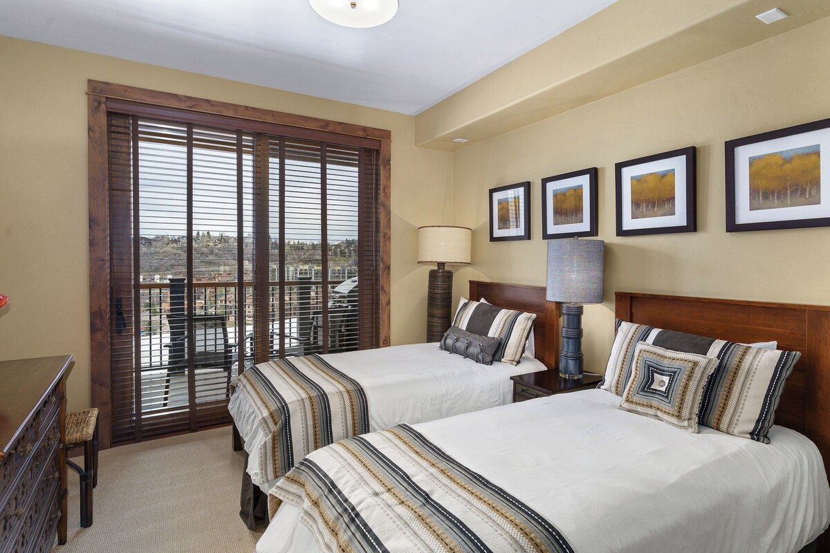 A hotel room with two beds, a desk, a chair, a lamp, and a view through a window with blinds.