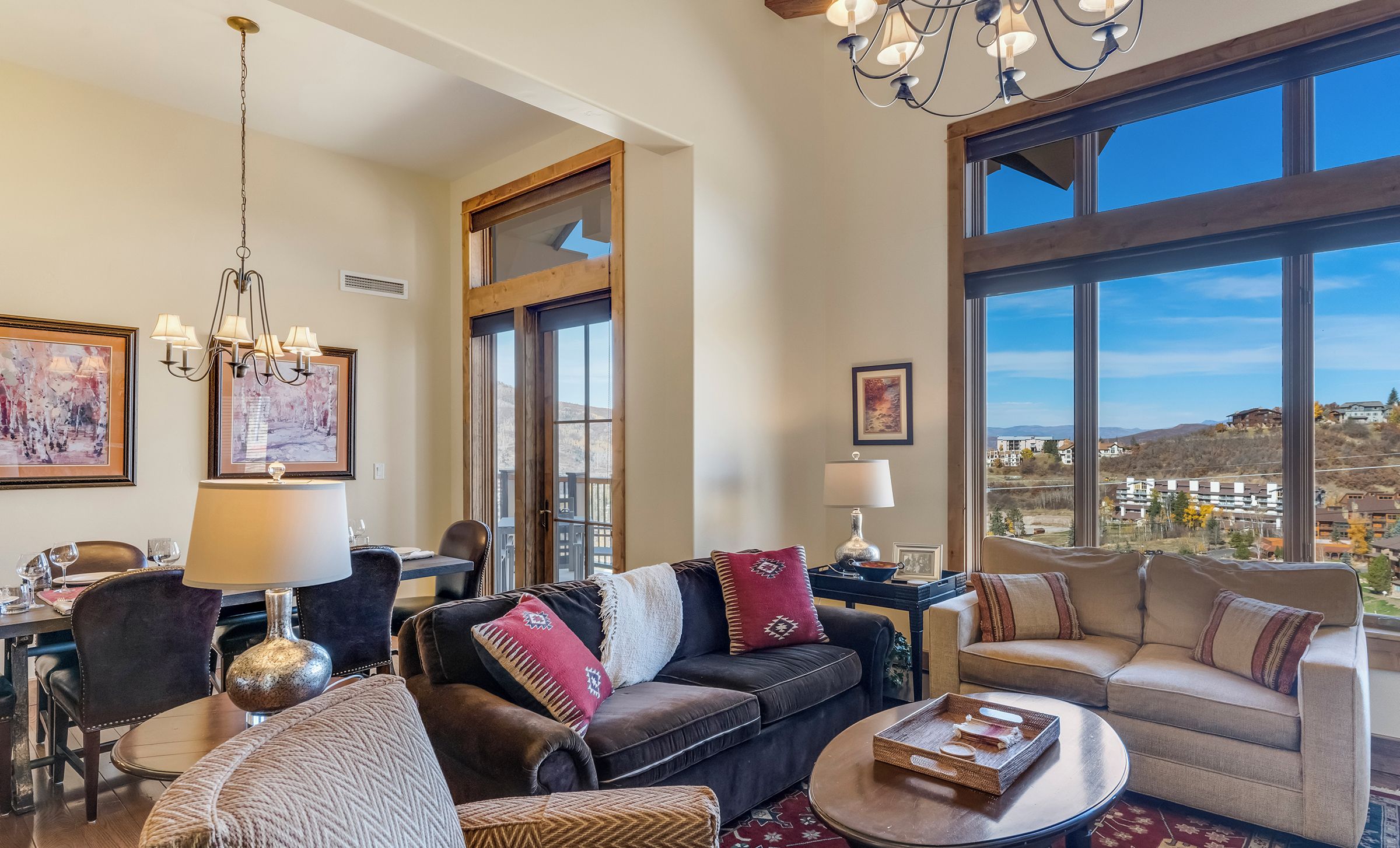 A spacious living room with a large window offering a view of a hillside. There are two sofas, a dining table, and a chandelier.