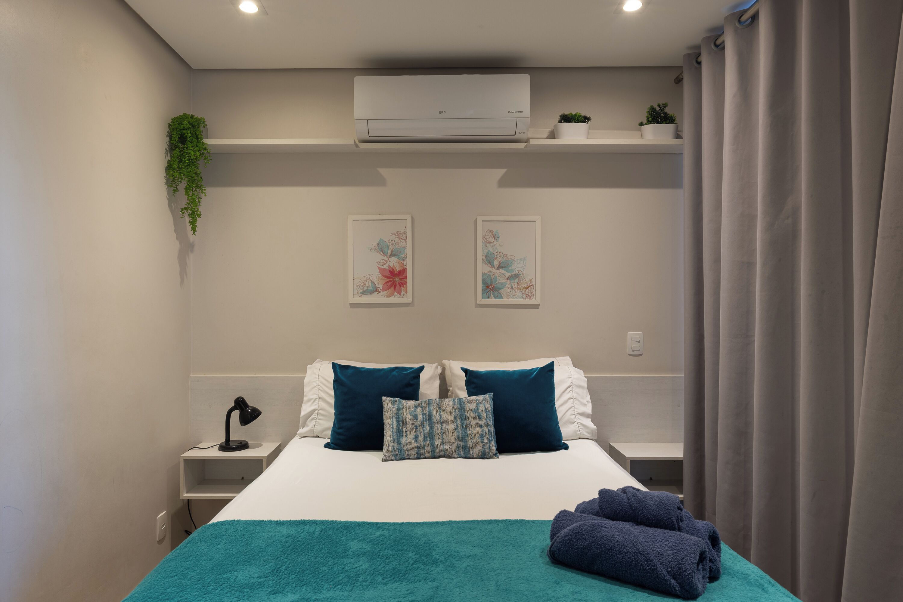 A hotel room with a bed, pillows, a bedside lamp, a small table, a plant, and artwork on the wall.