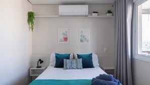 Un dormitorio moderno con cama, almohadas, lámpara de noche, ventana con cortinas, equipo de aire acondicionado y dos obras de arte colgadas en la pared.