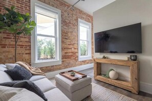 A living room with a brick wall, large windows, a flat-screen TV, a wooden console table, and a sofa with cushions. - Chestnut Chateau at the Brent Lofts (Pensacola)