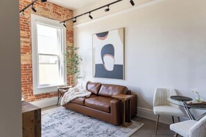 A living room with a brown leather sofa, a white armchair, a glass coffee table, and a large abstract painting on the wall. - Pura Vida at The Brent Lofts (Pensacola)