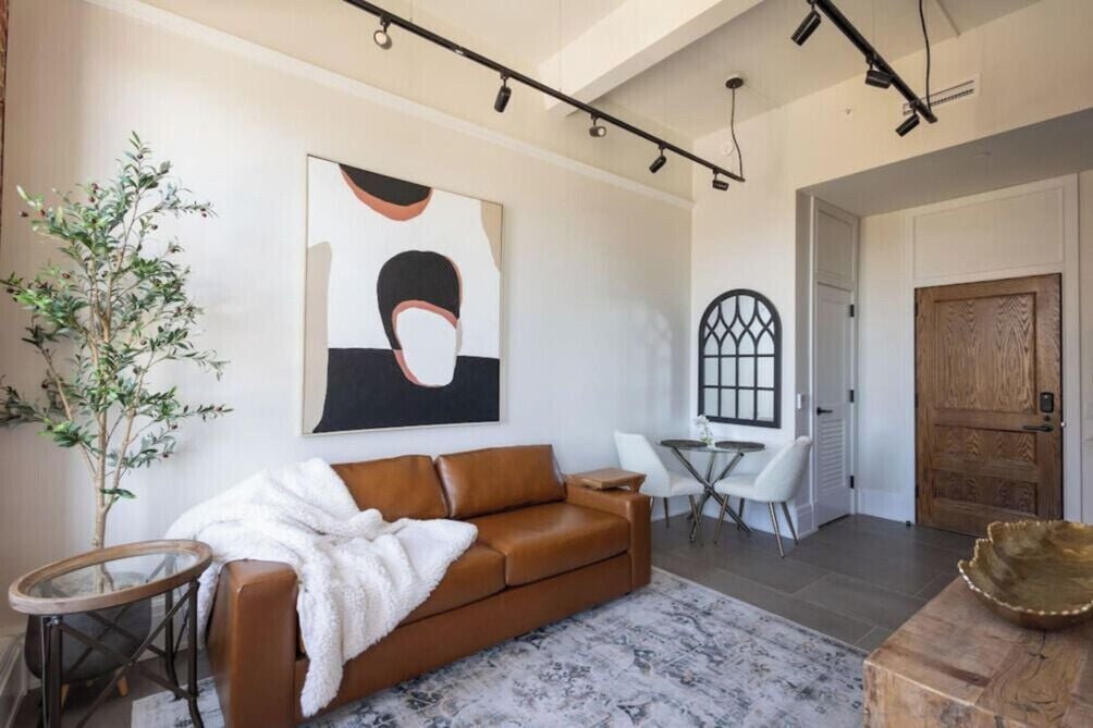 A modern living room with a brown leather sofa, a white throw blanket, a round coffee table, a glass side table, a potted plant, a large abstract painting, a wooden door, and a small round table with chairs.