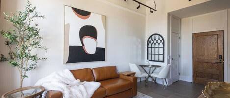 A modern living room with a brown leather sofa, a white throw blanket, a round coffee table, a glass side table, a potted plant, a large abstract painting, a wooden door, and a small round table with chairs.