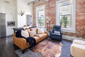 A modern living space with a leather sofa, a blue armchair, and a rustic brick wall. - The Brent Lofts Southern Comfort (Pensacola)