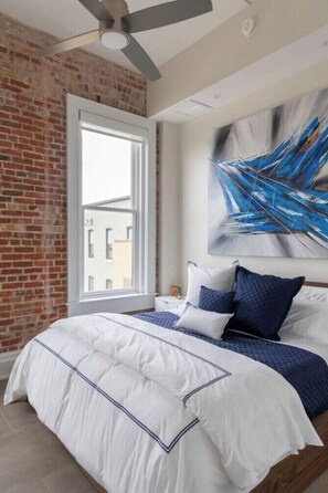Ein Schlafzimmer mit Backsteinwand, einem großen Fenster, einem Deckenventilator und einem modernen, abstrakten Gemälde.