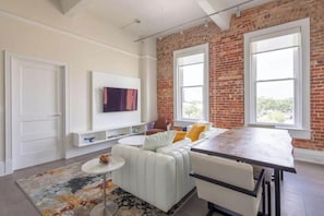 A modern living space with a brick wall, a white sofa, a dining table, and a TV mounted on the wall. - Modern Loft, Historic Streets- 2 bed/2 bath (Pensacola)