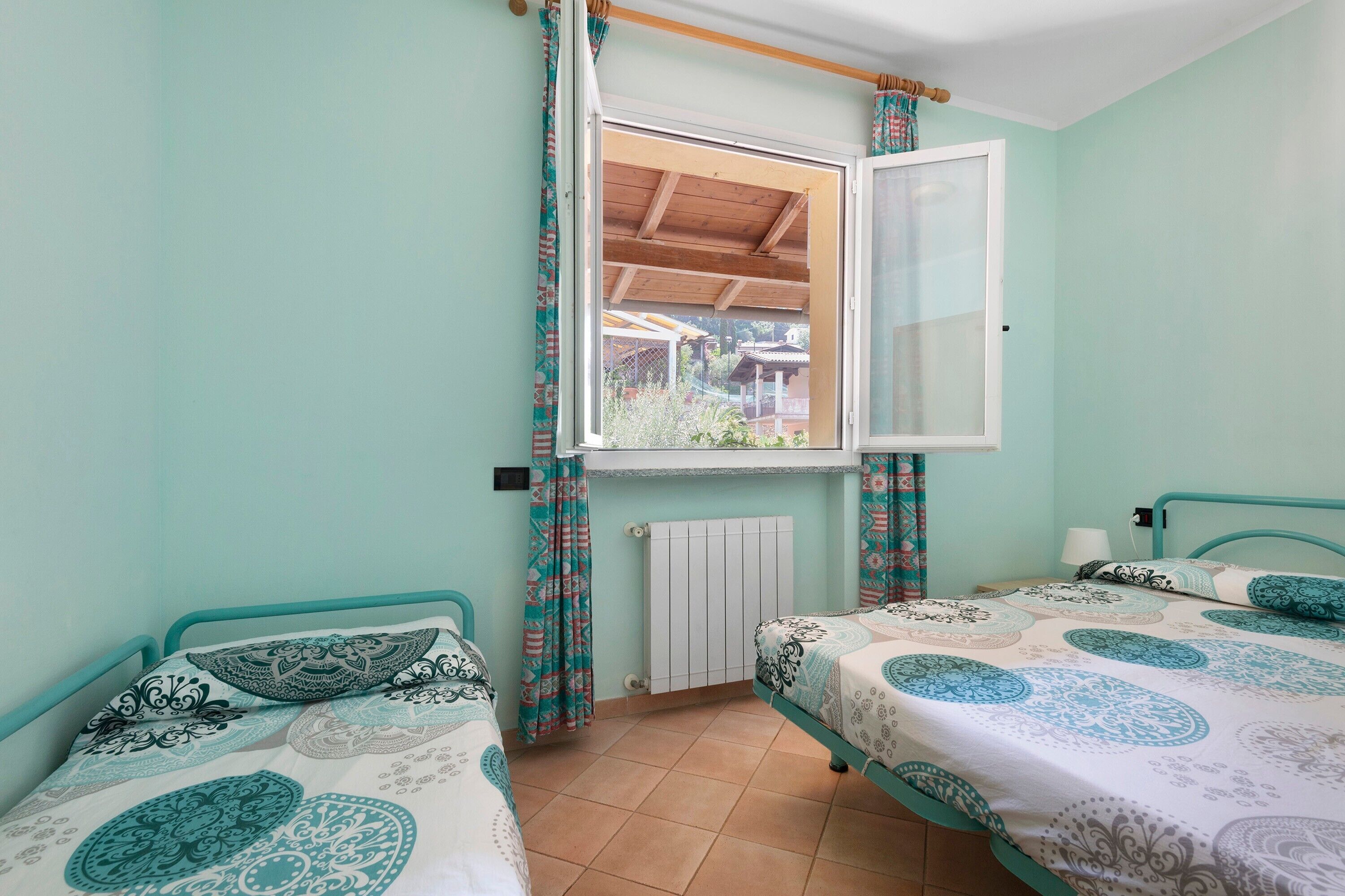 Une chambre avec deux lits, une fenêtre avec des rideaux, un radiateur et une vue sur une maison à l’extérieur.