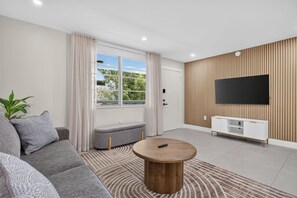 A modern living room with a sofa, ottoman, coffee table, and a TV on a wooden wall. - Cozy-Luxe Jr 1BR Apt | Freshly Remodeled & Stylish (Miami Beach)