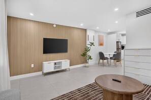 A modern living room with a flat-screen TV, a white entertainment unit, a dining area with a table and chairs, and a kitchen area with cabinets. - Cozy-Luxe Jr 1BR Apt | Freshly Remodeled & Stylish (Miami Beach)