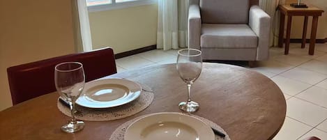 Una mesa de comedor con platos y vasos, un sofá, una lámpara y vistas a edificios a través de amplios ventanales.