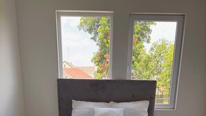 Une chambre à coucher avec un lit, deux fenêtres et une vue sur les arbres et les immeubles.