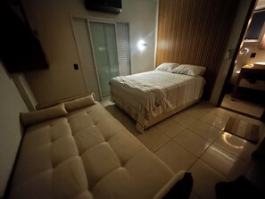 A bedroom with a bed, a sofa, a television mounted on the wall, and a wooden panel on the wall. - Modern retreat with barbecue, 200m from the beach. (Praia Grande)