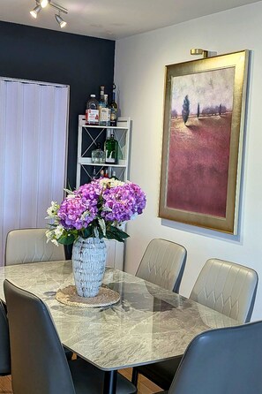 En matplats med ett bord med glasskiva, fyra stolar, en vas med blommor och en hylla med flaskor.