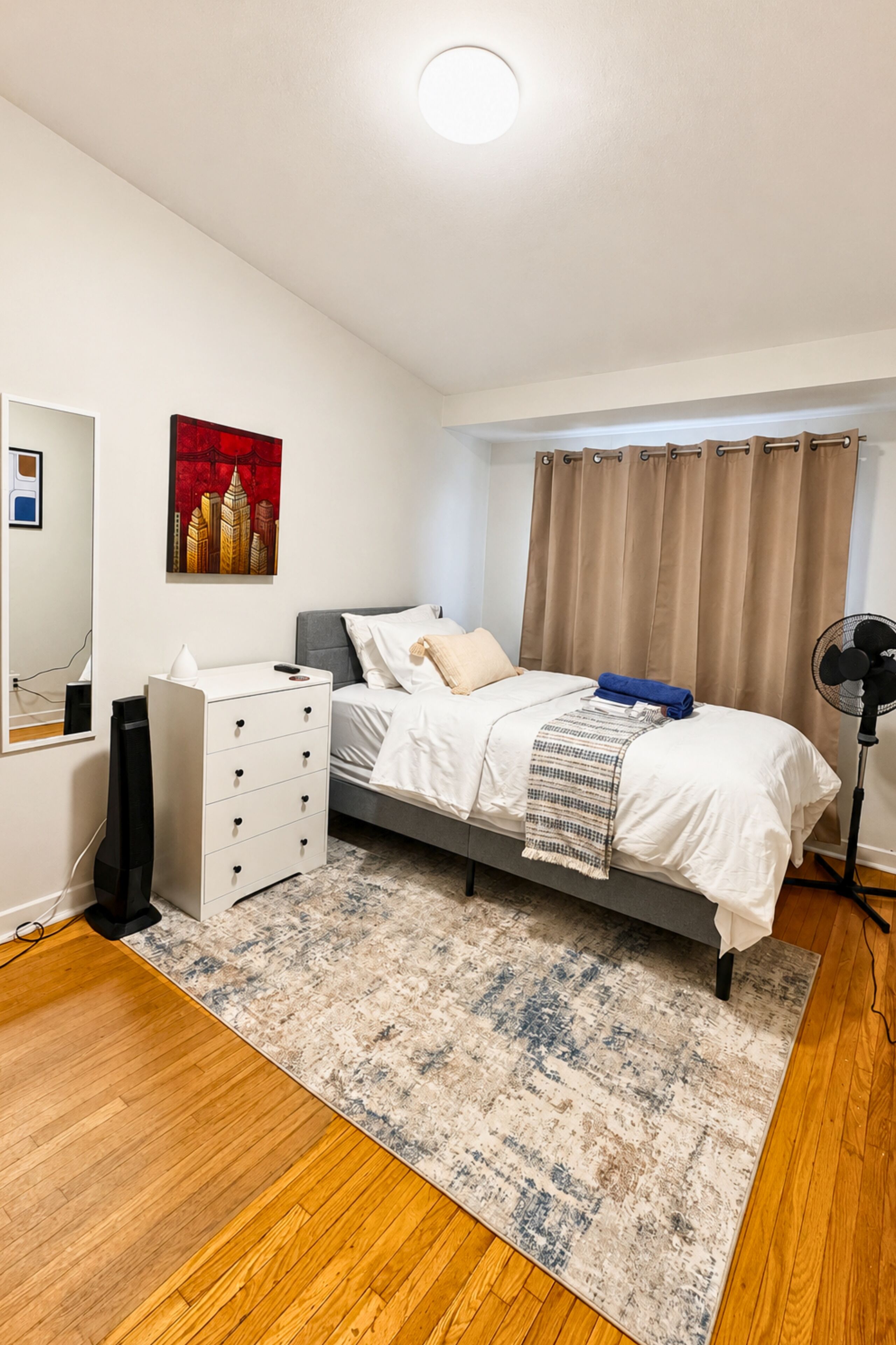 A bedroom with a bed, a dresser, a mirror, a painting, and a fan.
