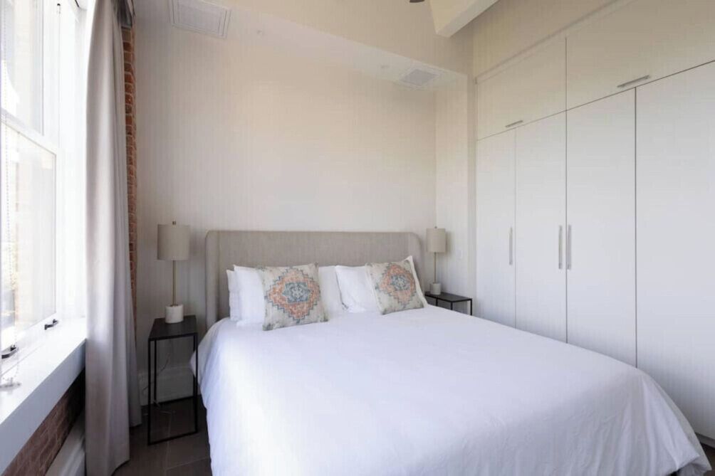 A bedroom with a bed, two bedside tables with lamps, a window with curtains, and a wardrobe.