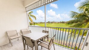 A balcony with a table and chairs, a view of a golf course, and a palm tree. - The Fairway Retreat at GreenLinks (Naples)
