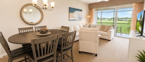 A dining area with a round table and chairs, a chandelier, and a large mirror. There is a sofa and a TV in the living area with a view of a green landscape.
