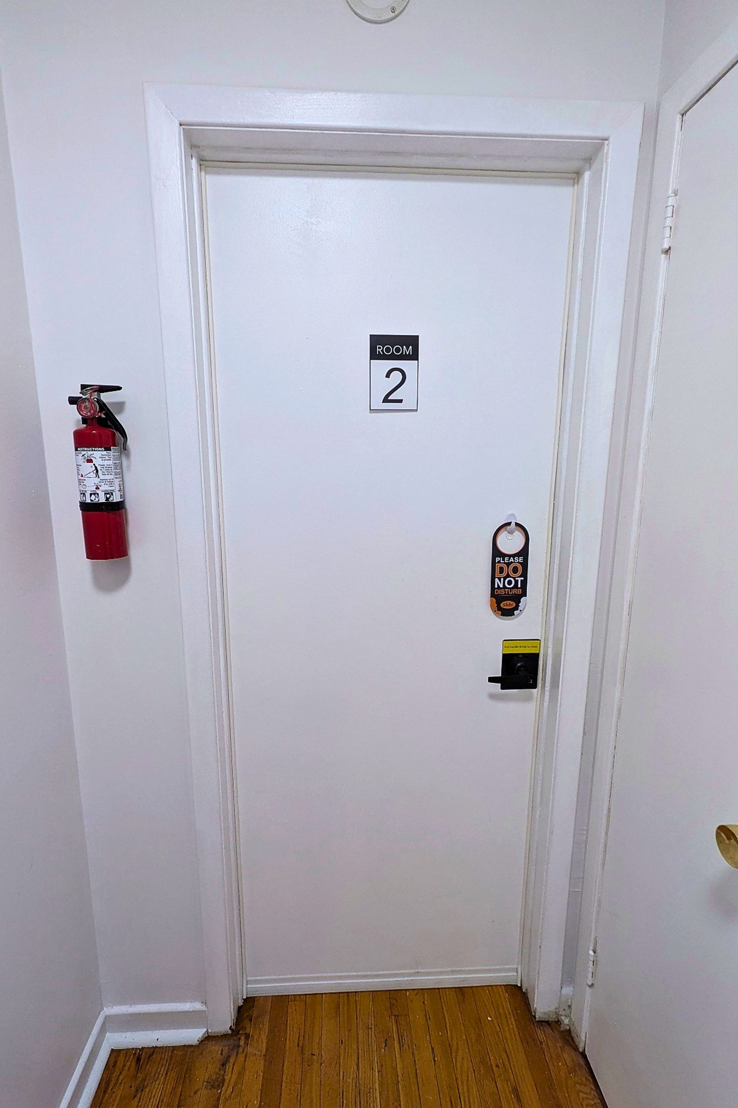 A white door with a room number sign, a fire extinguisher, and a doorbell.