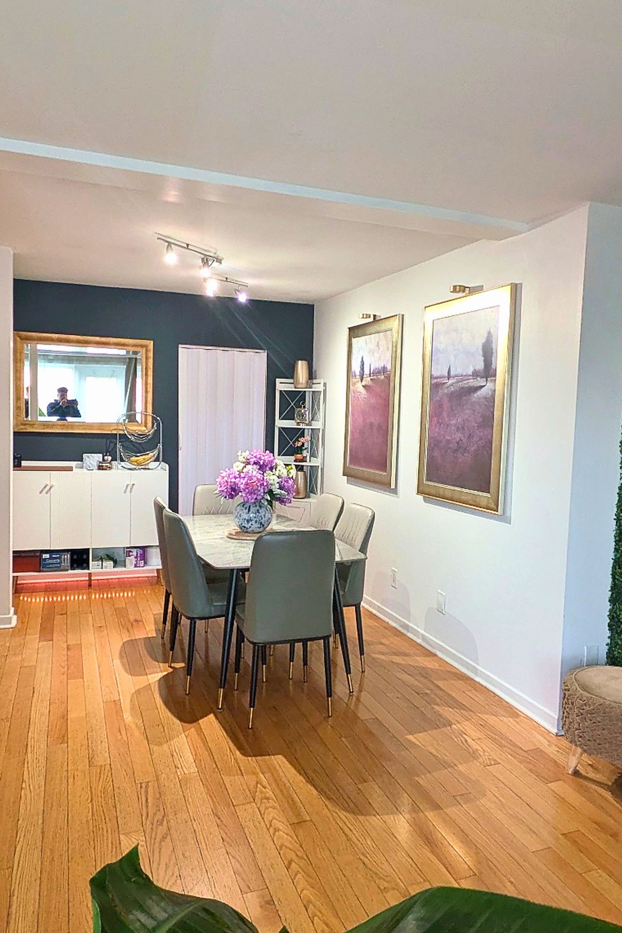 Un coin repas avec une table, des chaises et un vase de fleurs. Des tableaux encadrés ornent le mur, et un miroir est placé au-dessus de la table.