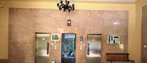 Vestíbulo con piso de mármol, dos ascensores, un banco y una araña.