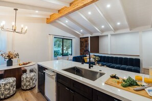 A modern kitchen with a marble countertop, a built-in dishwasher, and a dark blue sofa in the background. - Luxury Wellness Retreat with Cedar Sauna, Cold Plunge, Gym and Outdoor Oasis (Los Angeles)