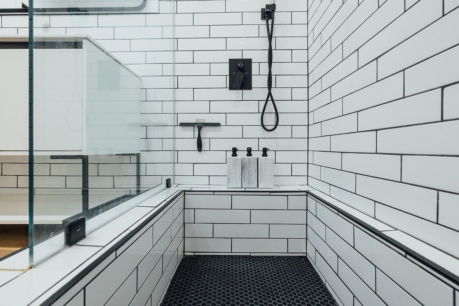 Une salle de bain moderne avec des carreaux de métro blancs, un drain de douche noir et trois flacons de shampoing, d’après-shampoing et de gel douche.
