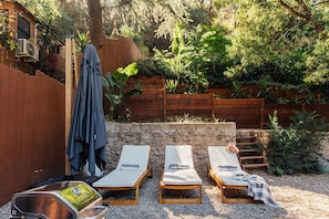 A backyard with a wooden fence, a stone wall, and three lounge chairs with cushions. - Luxury Wellness Retreat with Cedar Sauna, Cold Plunge, Gym and Outdoor Oasis (Los Angeles)