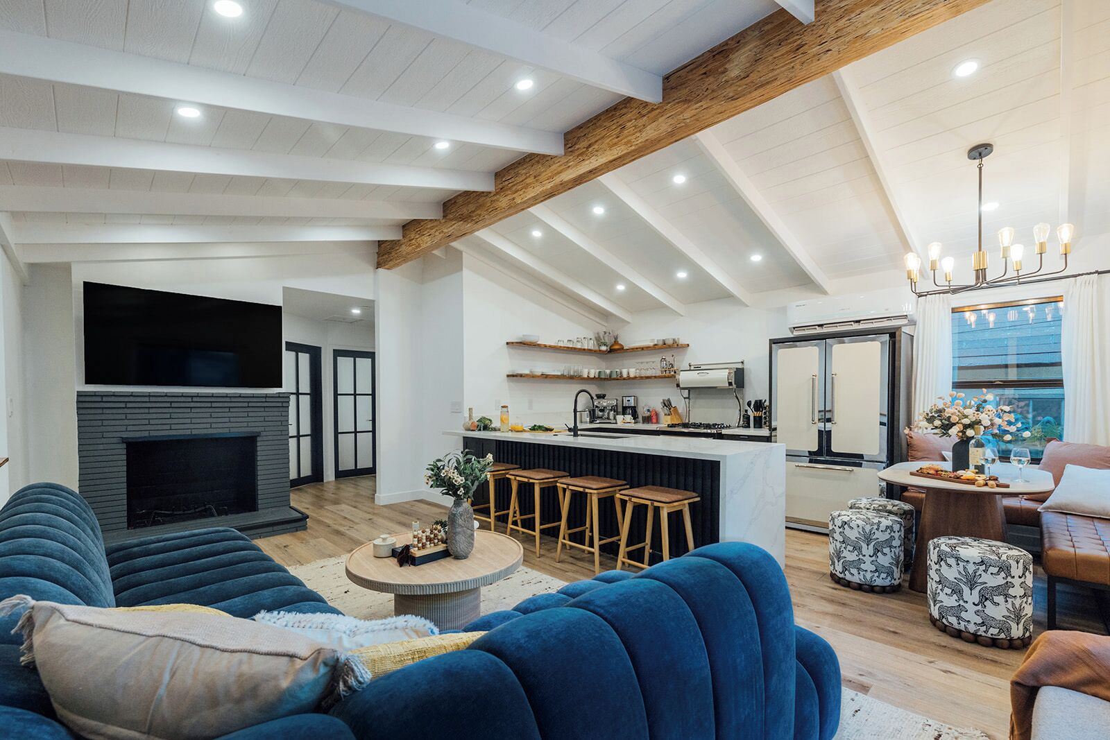 A modern living room with a fireplace, blue sofas, a kitchen with a bar area, and a dining area with a chandelier.