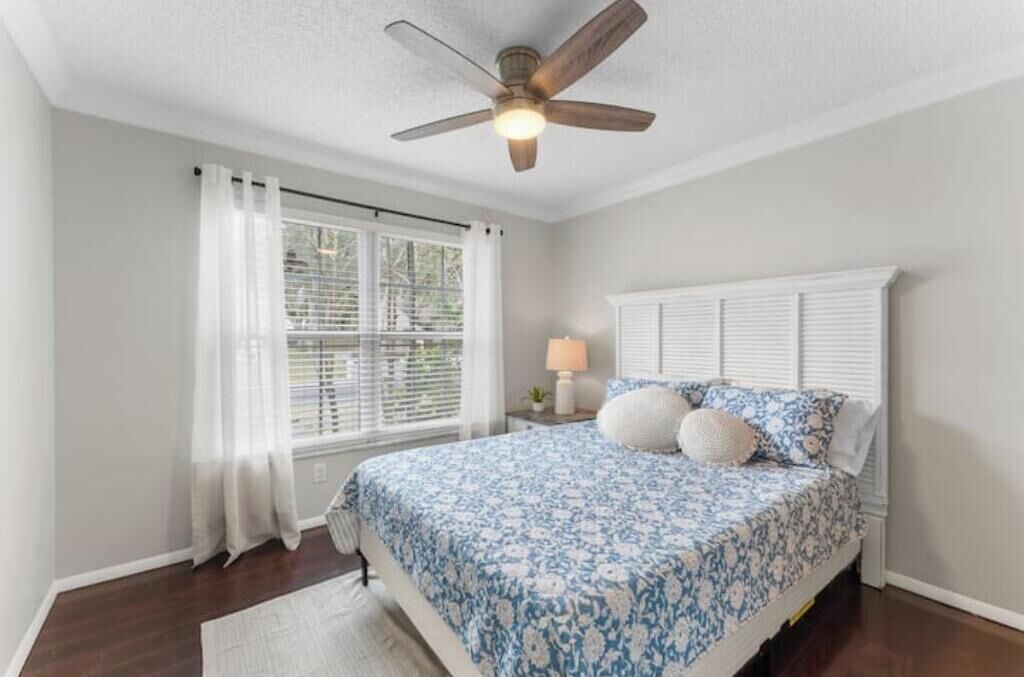 A bedroom with a bed, a ceiling fan, a window with curtains, and a nightstand with a lamp.